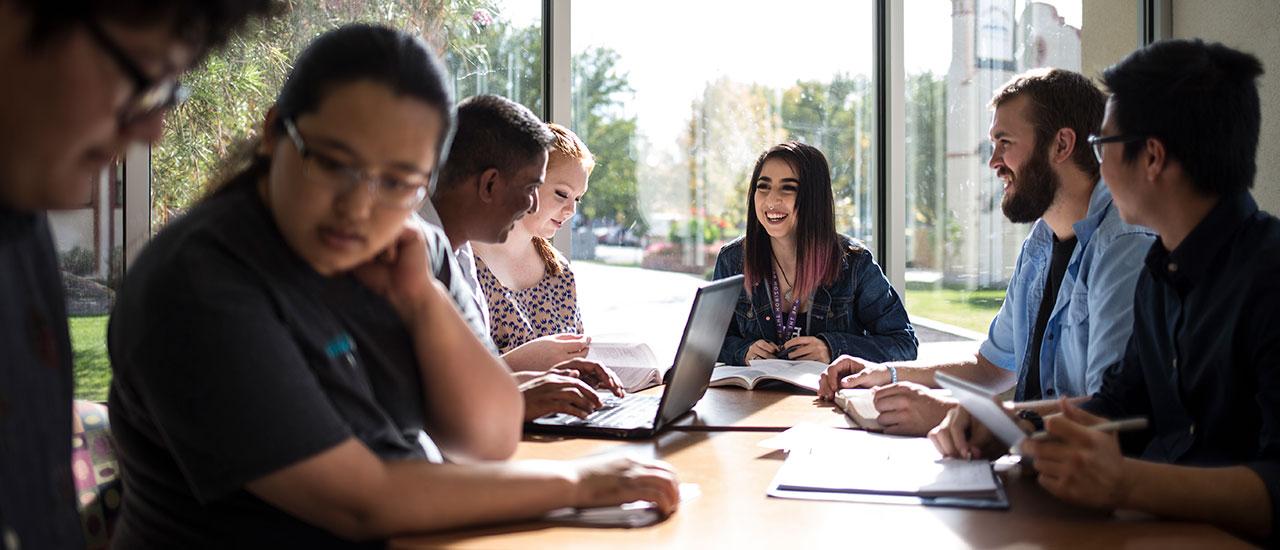 Students sitting at a table studiying together next to large windows. Outside of the windows is a view of greenery and buildings that are present on campus.